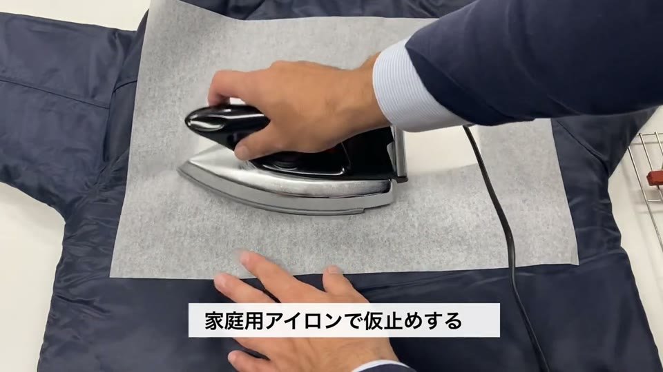 紺色のジャケットにのせたマークの上に当て紙を置き、家庭用アイロンで押さえて仮止めする様子と、家庭用アイロンで仮止めするというテロップ