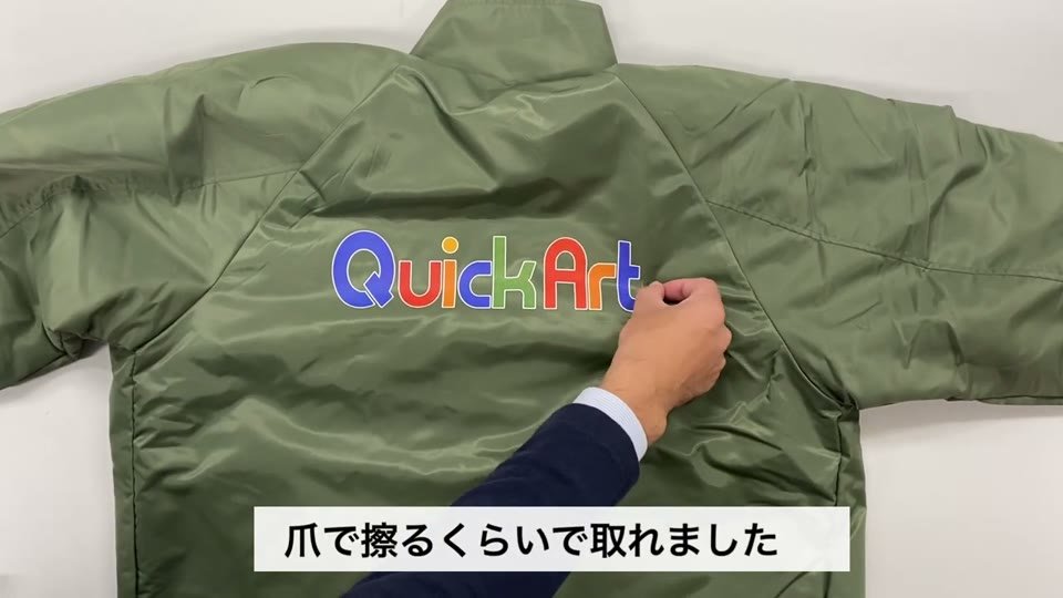 カーキ色のナイロンのコーチジャケットの背中に、カラーのQuickArtのロゴがプリントされ、指でロゴの端をこすっている様子と、爪で擦るくらいで取れましたというテロップ