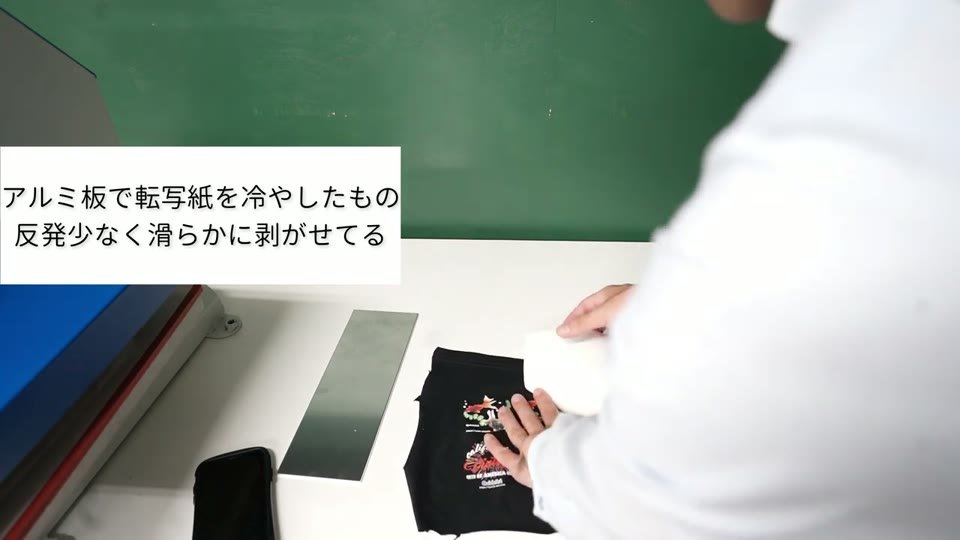 机の上にアルミ板と黒いTシャツ。アルミ板で冷やしたTシャツから転写紙を剥がしている。テロップに「アルミ板で転写紙を冷やしたもの 反発少なく滑らかに剥がせてる」