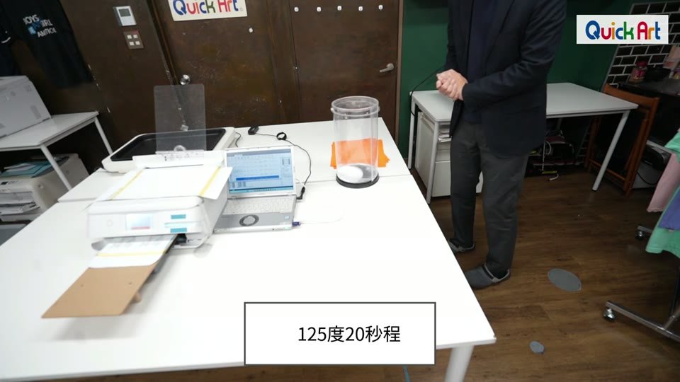 ショールームの作業台で小型DTFプリンターが印刷を続け、奥の熱プレス機でプレスしている様子。125度20秒程というテロップ