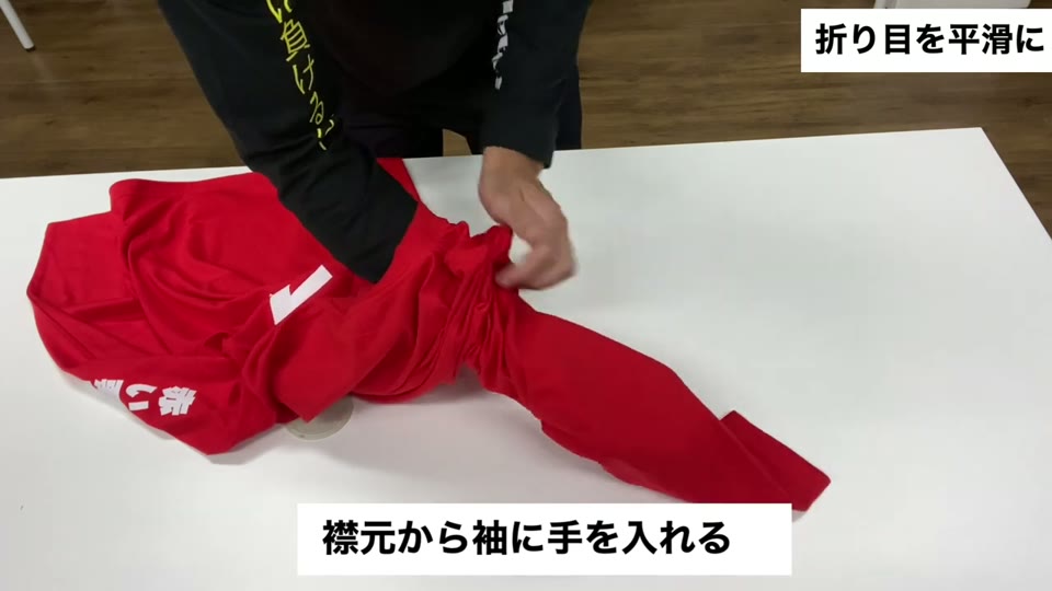 赤いロンTの襟元から袖の中に手を入れて折り目を整えている様子。襟元から袖に手を入れるとテロップが出ている