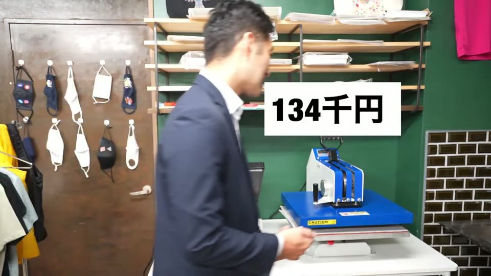 ショールームに並ぶ2台の熱プレス機のうち、青いスイング式のプレス機の前で、134千円というテロップ