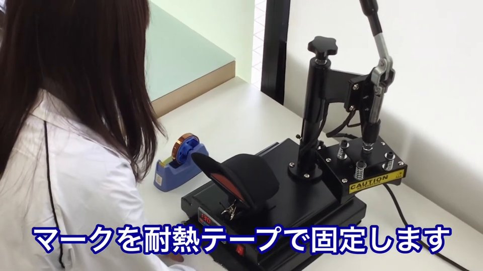 帽子プレス機に黒いキャップをセットし、マークを留めている様子。テロップ:マークを耐熱テープで固定します