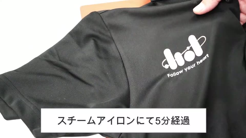 スチームアイロンをかけて5分たった黒いポロシャツの胸のプリント。プリントのまわりの四角いテカリがほとんど見えなくなっている様子と、スチームアイロンにて5分経過というテロップ