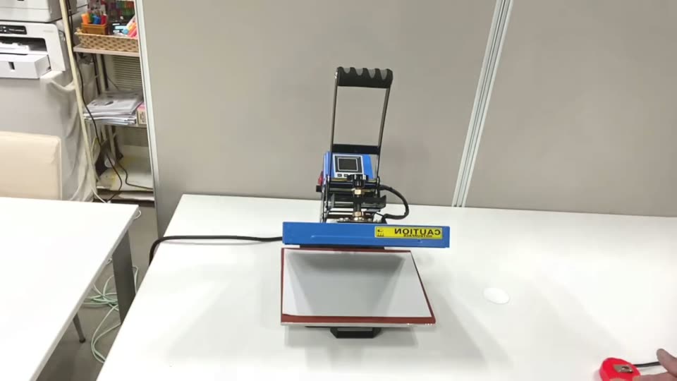 白い作業台に置いた平板あおり式ミニプレス機 JL-PL001E。青い上ゴテと赤いラバー面の下ゴテ