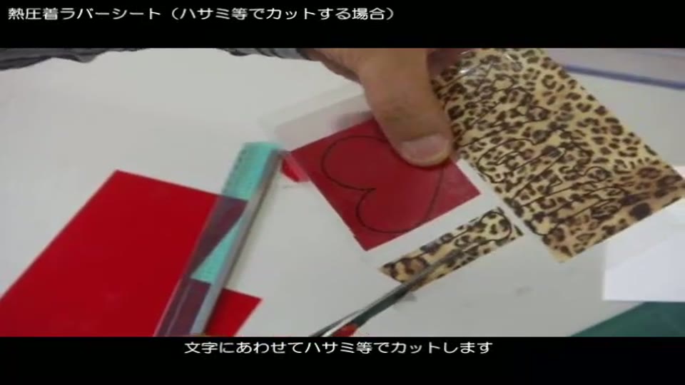 透明フィルムを貼った赤いラバーシートと柄入りラバーシートを、赤い柄のハサミで文字の形に切っているところ