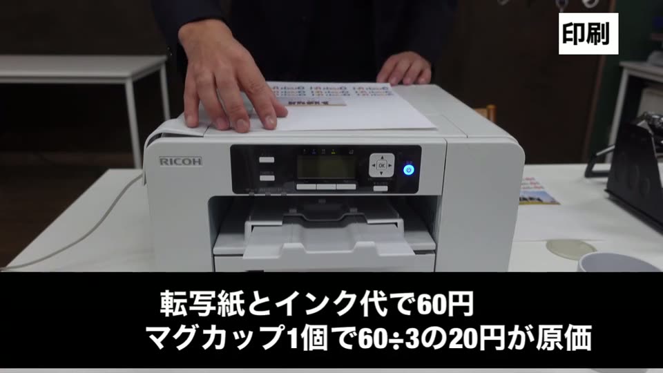 昇華プリンターで印刷する手元。転写紙とインク代で60円、マグカップ1個で60割る3の20円が原価というテロップ