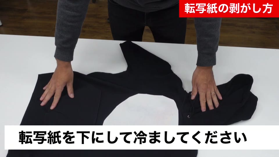 黒いパーカーを裏返し、転写紙を下にして作業台に置いている様子。転写紙を下にして冷ましてくださいとテロップが出ている