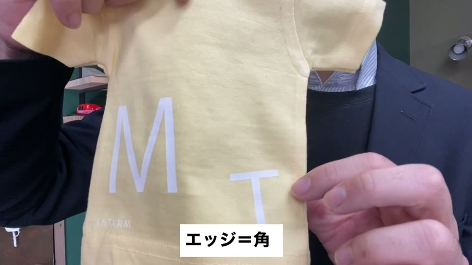 黄色いミニTシャツにプリントした白いMとTの文字の角を指さし、エッジ＝角と字幕が出ている様子