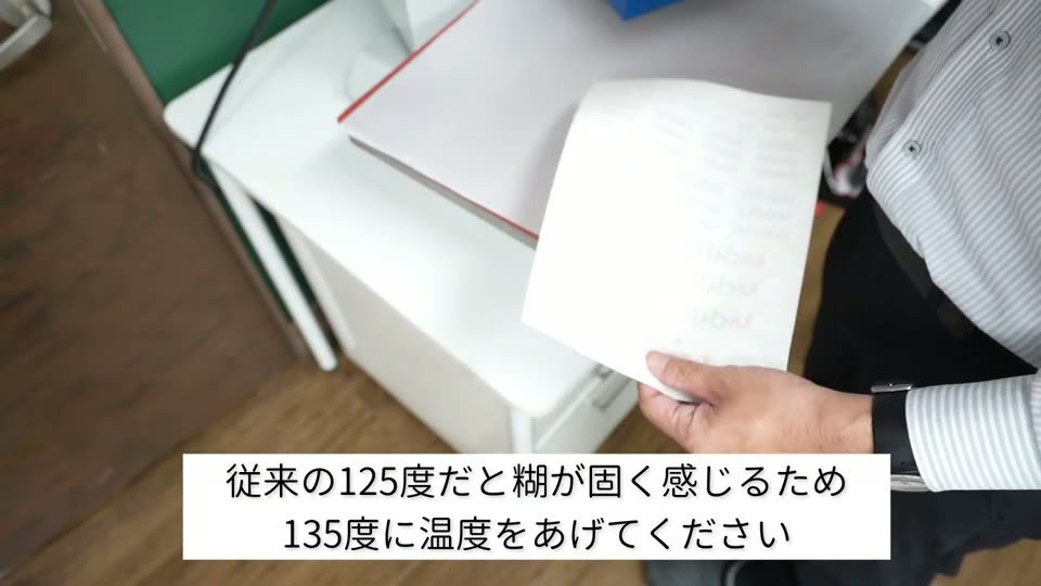 プレス機の前で剥がしたシートを持っている。テロップは従来の125度だと糊が固く感じるため135度に温度をあげてください