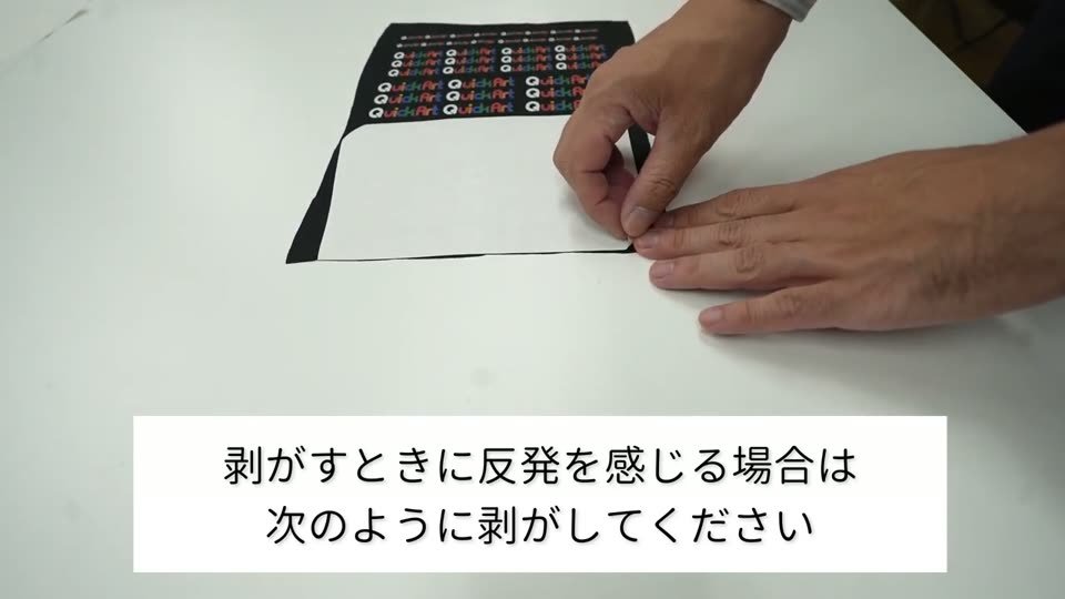 黒い生地を左手で押さえ、右手で裏紙を低く折り返しながら少しずつ剥がしている。テロップは剥がすときに反発を感じる場合は次のように剥がしてください