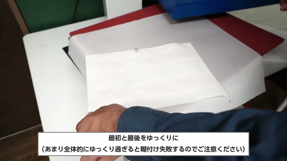 プレス台の上で糊シートを剥がし終えようとしている手元と、最初と最後をゆっくりに、あまり全体的にゆっくり過ぎると糊付け失敗するのでご注意くださいの字幕