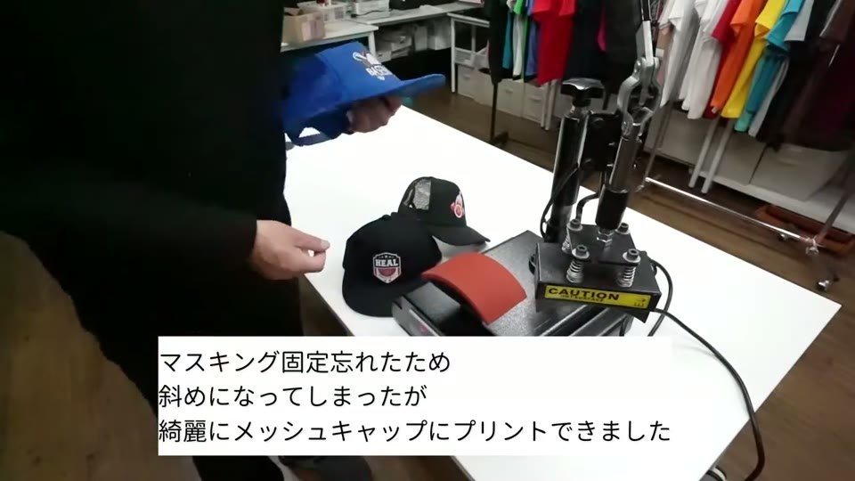 帽子プレス機の横で、白トナーでプリントした黒いメッシュキャップを見せている様子